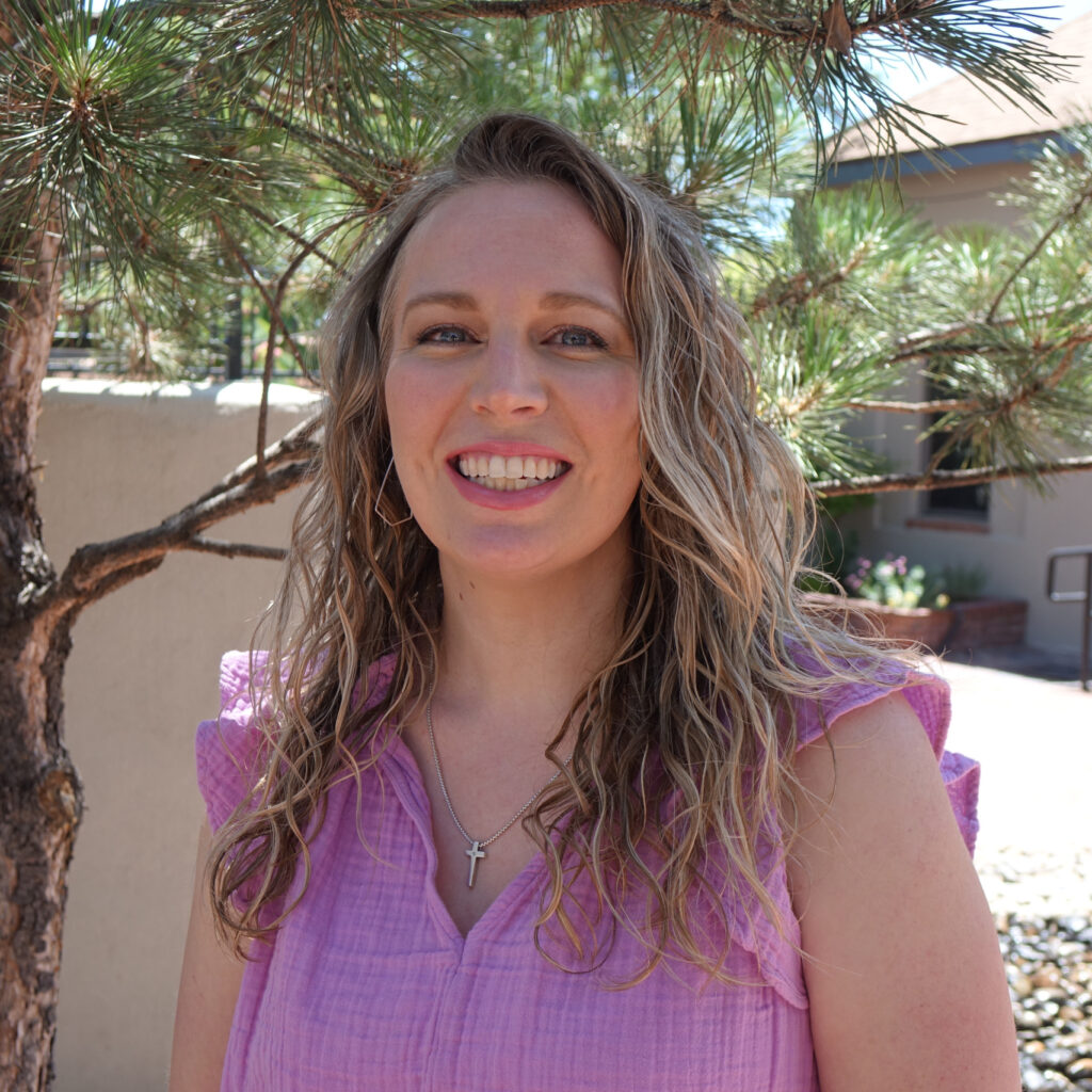 Ashley is smiling in front of a pine tree. She has long blonde hair and is wearing a purple blouse.